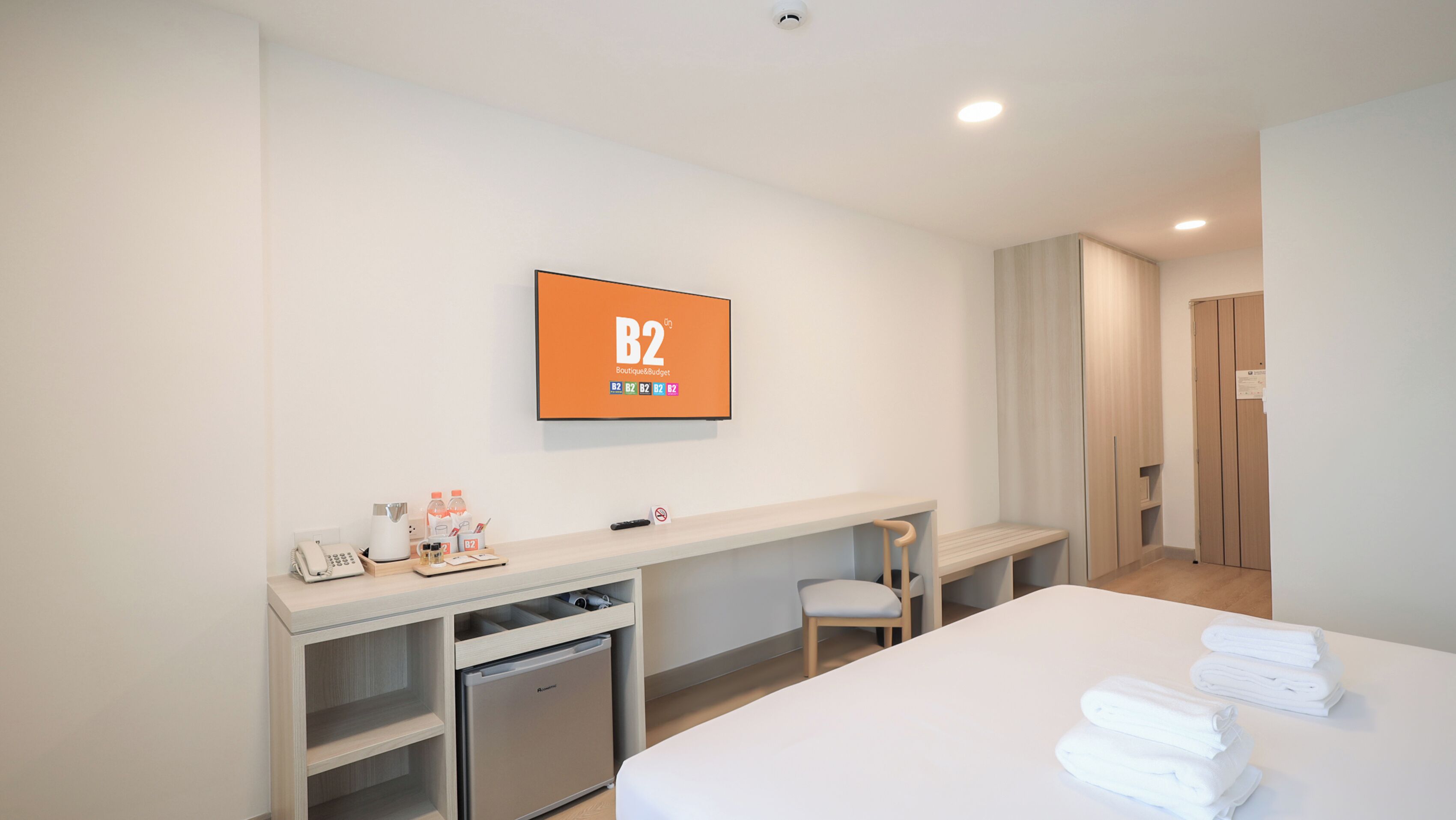 Une chambre d’hôtel équipée d’un lit, d’un bureau, d’un petit réfrigérateur, d’une chaise et d’un téléviseur affichant le message « B2 Boutique Budget ».