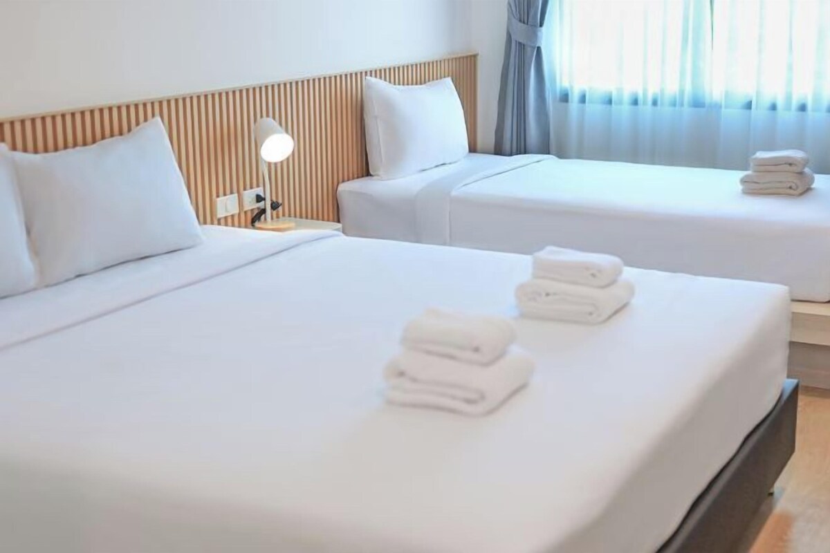 A hotel room with two beds, each with white linens and pillows, a bedside lamp, and a window with curtains.