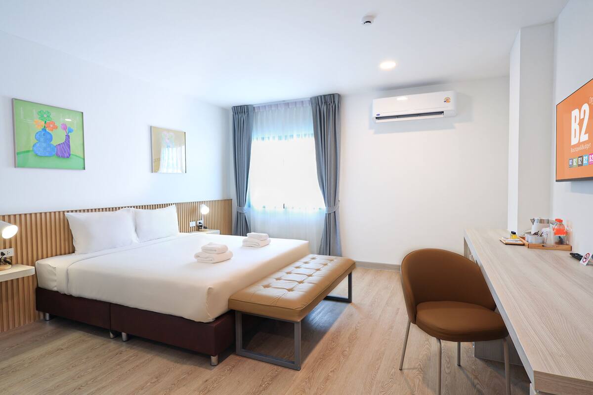 A hotel room with a bed, bedside table, chair, desk, TV, air conditioning unit, and artwork on the walls.