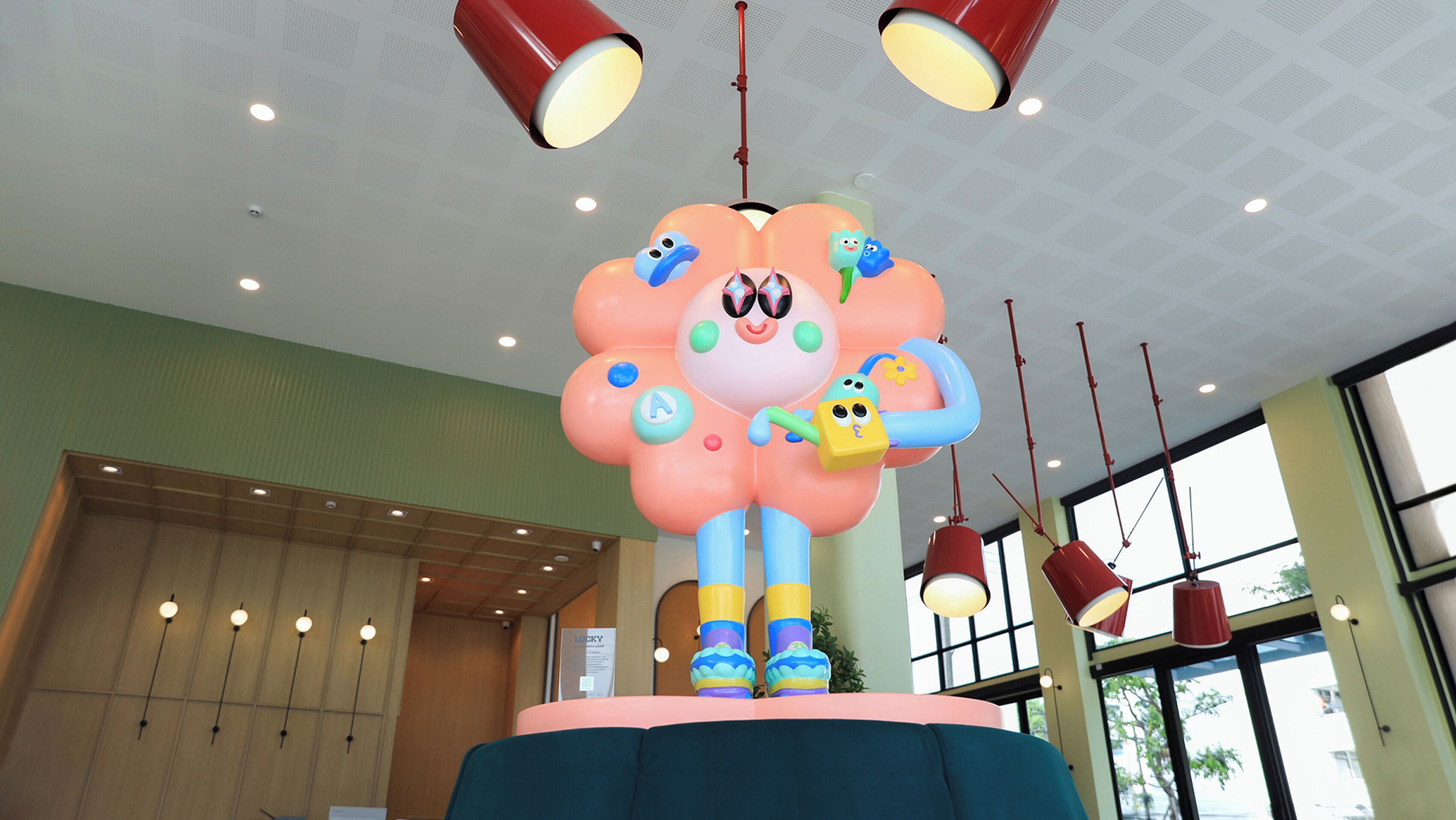 Un'ampia scultura colorata in stile cartoon raffigura un personaggio dalla testa a forma di fiore, posizionato su una piattaforma in una stanza con rivestimenti in legno e lampade rosse pendenti.
