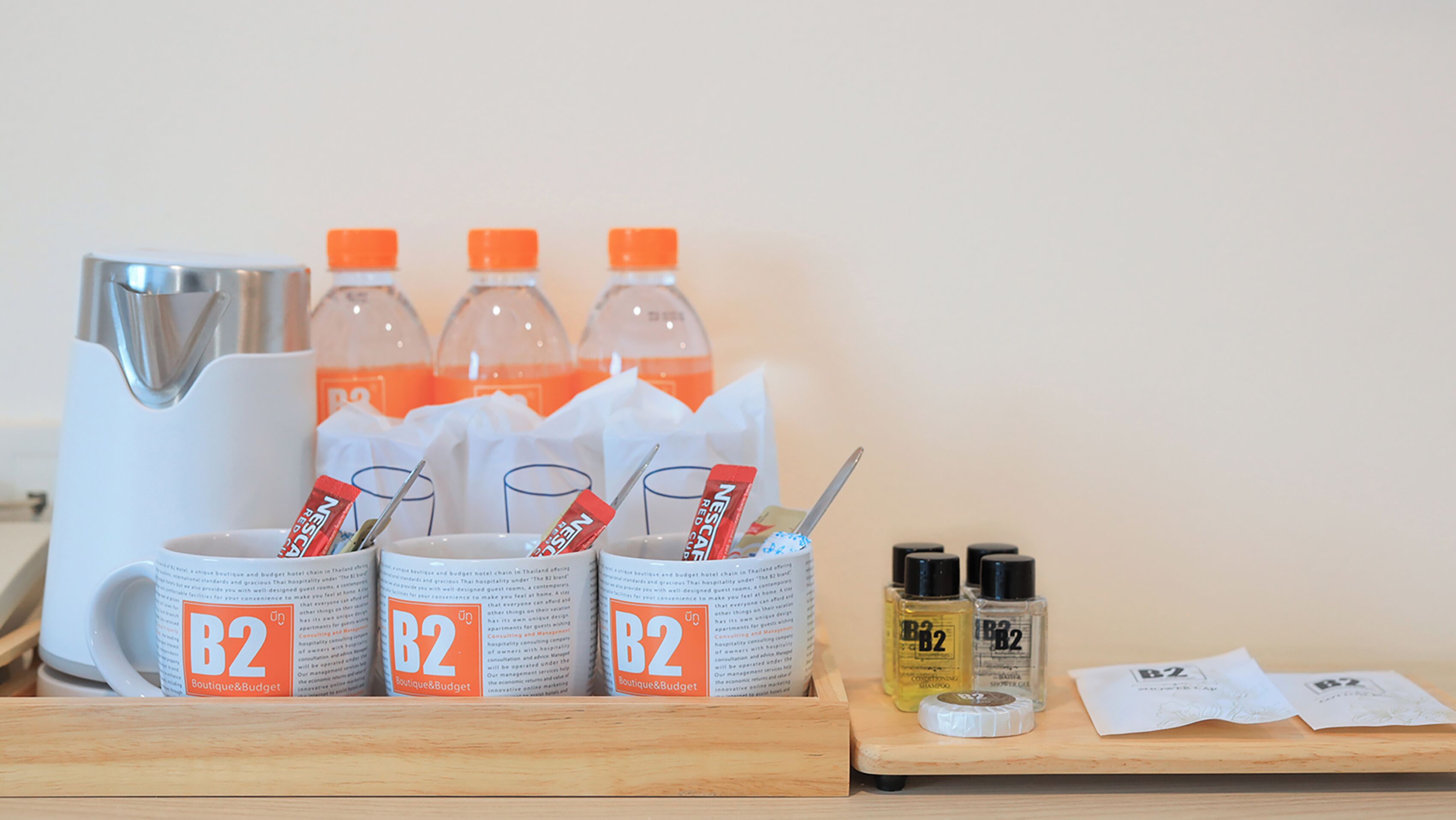 A hotel room setup with a coffee maker, three bottles of orange juice, two mugs with B2 Boutique&Budget branding, a small tray with toiletries including shampoo and shower gel, and a tissue box.