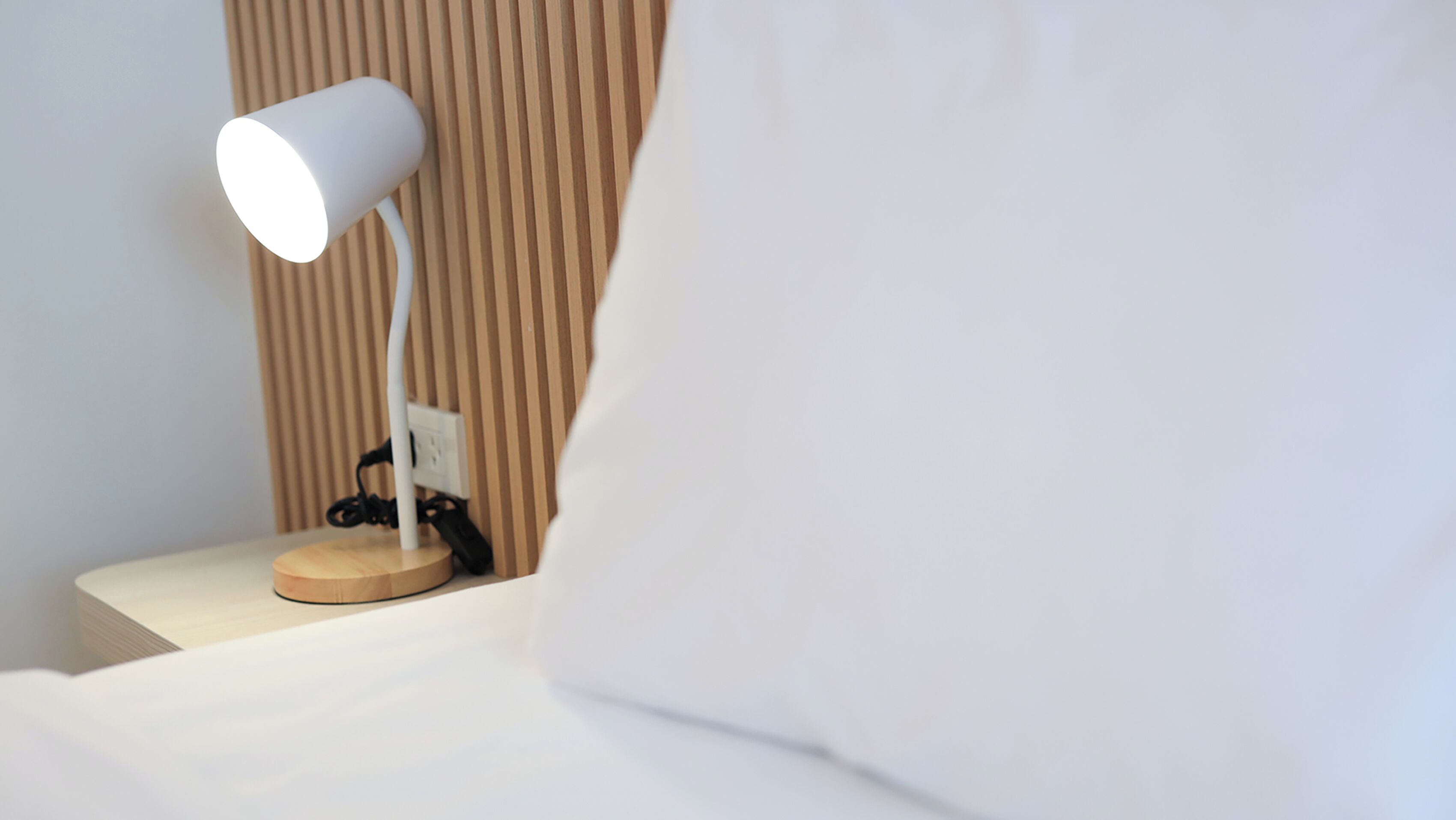 Een nachtkastje met een witte lamp, een stopcontact en een oplaadkabel.