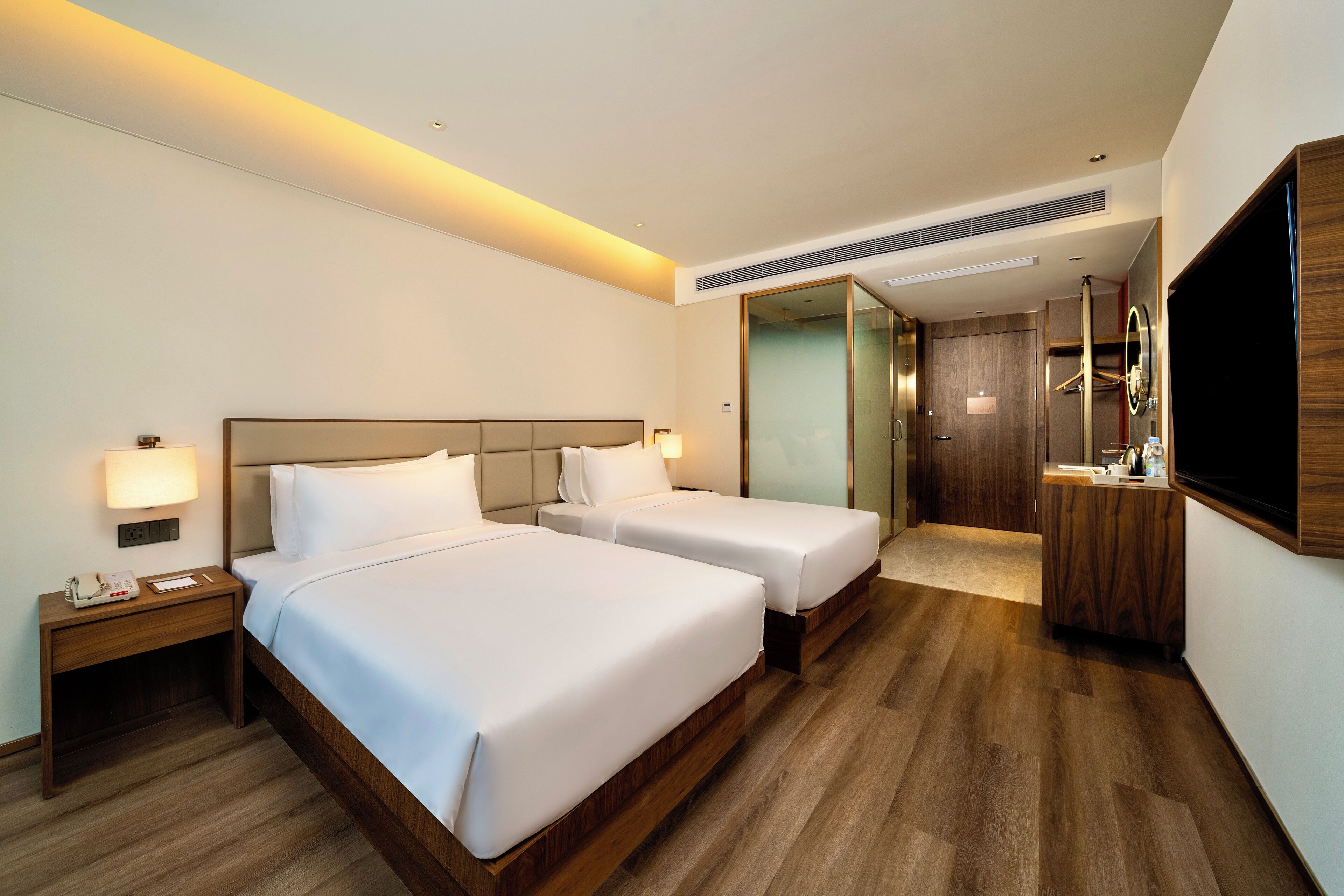 A hotel room with two beds, a bedside table with a lamp, a TV mounted on the wall, and a wooden floor.