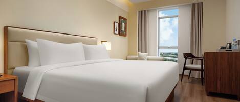 A hotel room with a large bed, a desk, a chair, and a view of the city through the window.