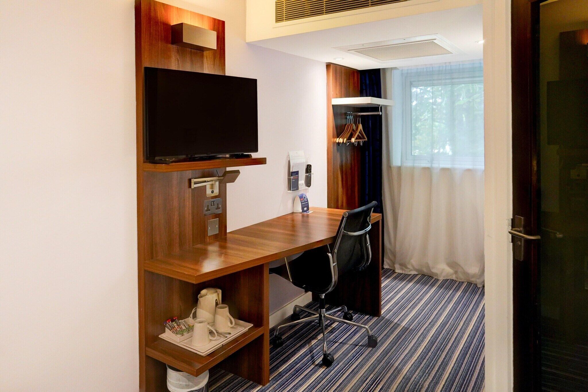 Habitación de hotel con escritorio de madera, televisor, perchero y ventana con cortinas.
