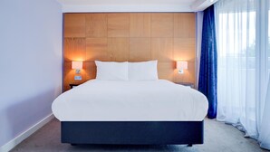 A modern hotel room with a large bed, wooden headboard, two bedside lamps, and sheer curtains.