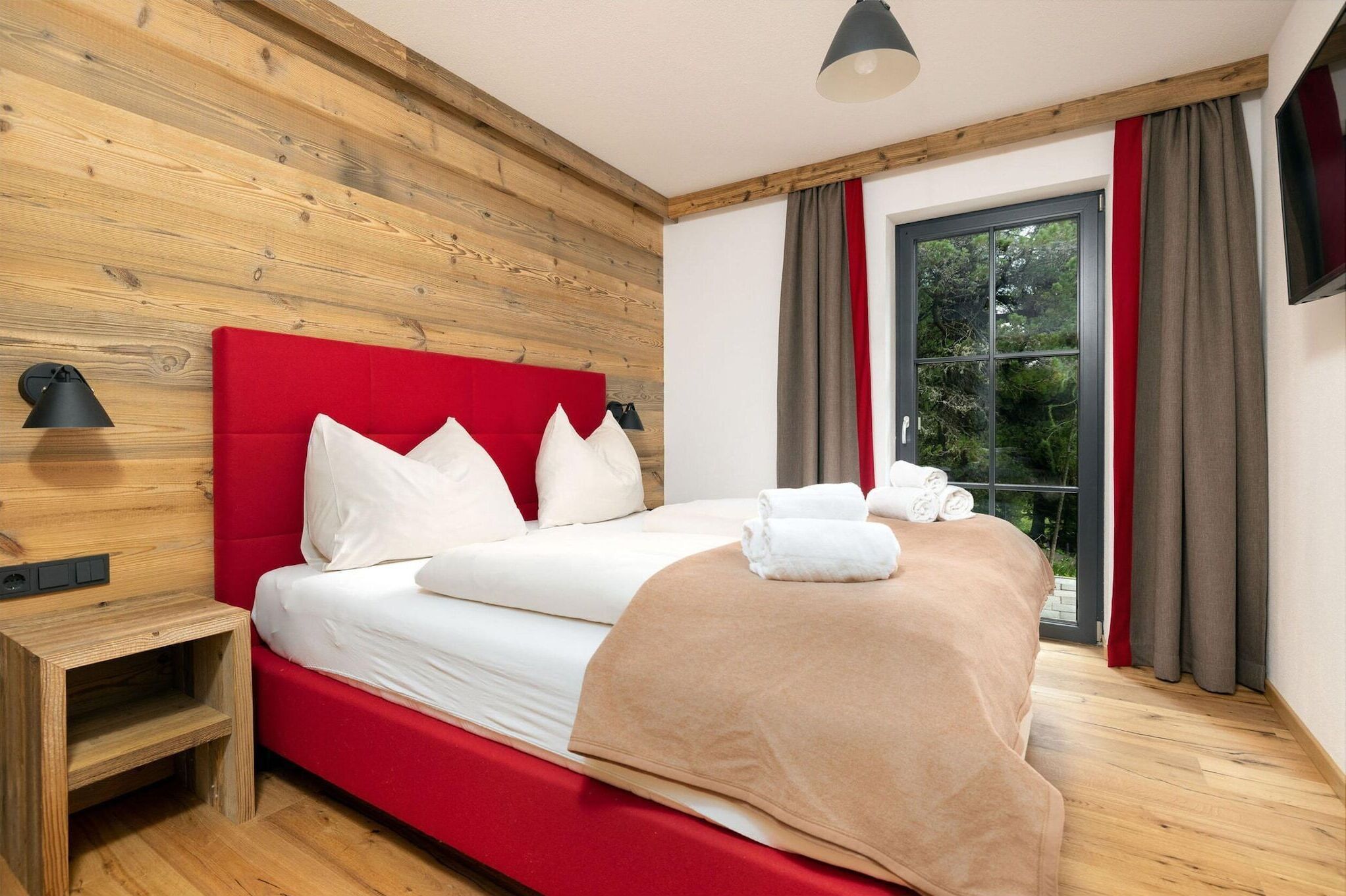 Habitación de hotel con pared de madera, cabecero rojo, ventana grande con cortinas, mesita de noche y cama con ropa blanca y manta color beige.