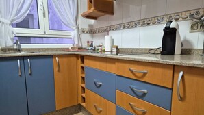 A kitchen with blue and wooden cabinets, a countertop, a coffee maker, and a window with curtains. - 25A06 Centrico Playa 4 dormitorios Luanco (Luanco)