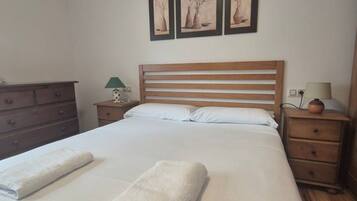 Cama bem arrumada com duas toalhas, cabeceira de madeira, mesa de cabeceira com abajur e cômoda.