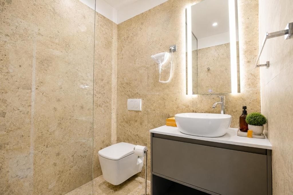 Un baño moderno con pared de mármol, inodoro blanco, lavamanos con espejo y una pequeña planta en maceta.