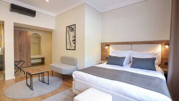 A modern bedroom with a large bed, a wooden headboard, a small table, a bench, and a wall-mounted artwork.