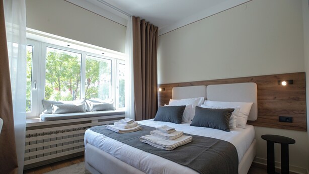 A modern bedroom with a large bed, a window seat, and a view of trees. - Simma Luxury Apartments & Rooms (Belgrade)