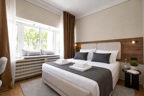 A modern hotel room with a large bed, a window with a view of trees, a bedside table with a bottle and a book, and a bench by the window. - Simma Luxury Apartments & Rooms (Belgrade)