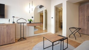 A modern living room with a wooden floor, a grey sofa, a coffee table, a TV mounted on the wall, a small plant on a side table, and a mirror reflecting the room. - Simma Luxury Apartments & Rooms (Belgrade)