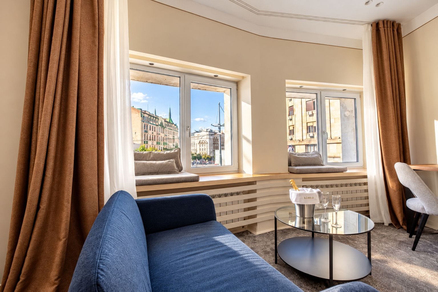 A modern hotel room with a sofa, a glass table, and a view of the cityscape through large windows.