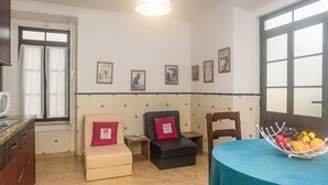 Ein gemütliches Zimmer mit einer Küchenzeile, zwei Stühlen, einem Tisch mit Obstschale und einem Fenster mit Vorhängen.