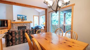 Een gezellig interieur van een chalet met een houten eettafel, stoelen, een stenen open haard, een tv aan de muur en een balkon met uitzicht op de natuur.