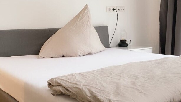 A neatly made bed with a white and beige duvet, a grey headboard, and a bedside table with a lamp and a power outlet.
