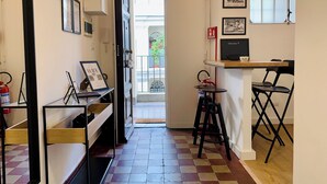 A narrow hallway with a checkered floor, leading to a door with a glass panel. There are framed pictures on the walls, a small table with a chair, and a fire extinguisher on the left.