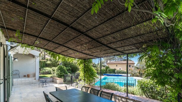 Um pátio externo com mesa e cadeiras, uma piscina ao fundo e uma área coberta com telhado de palha.