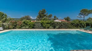 Piscina azul cristalina com borda azulejada, rodeada por um sebedo e árvores, sob um céu limpo.