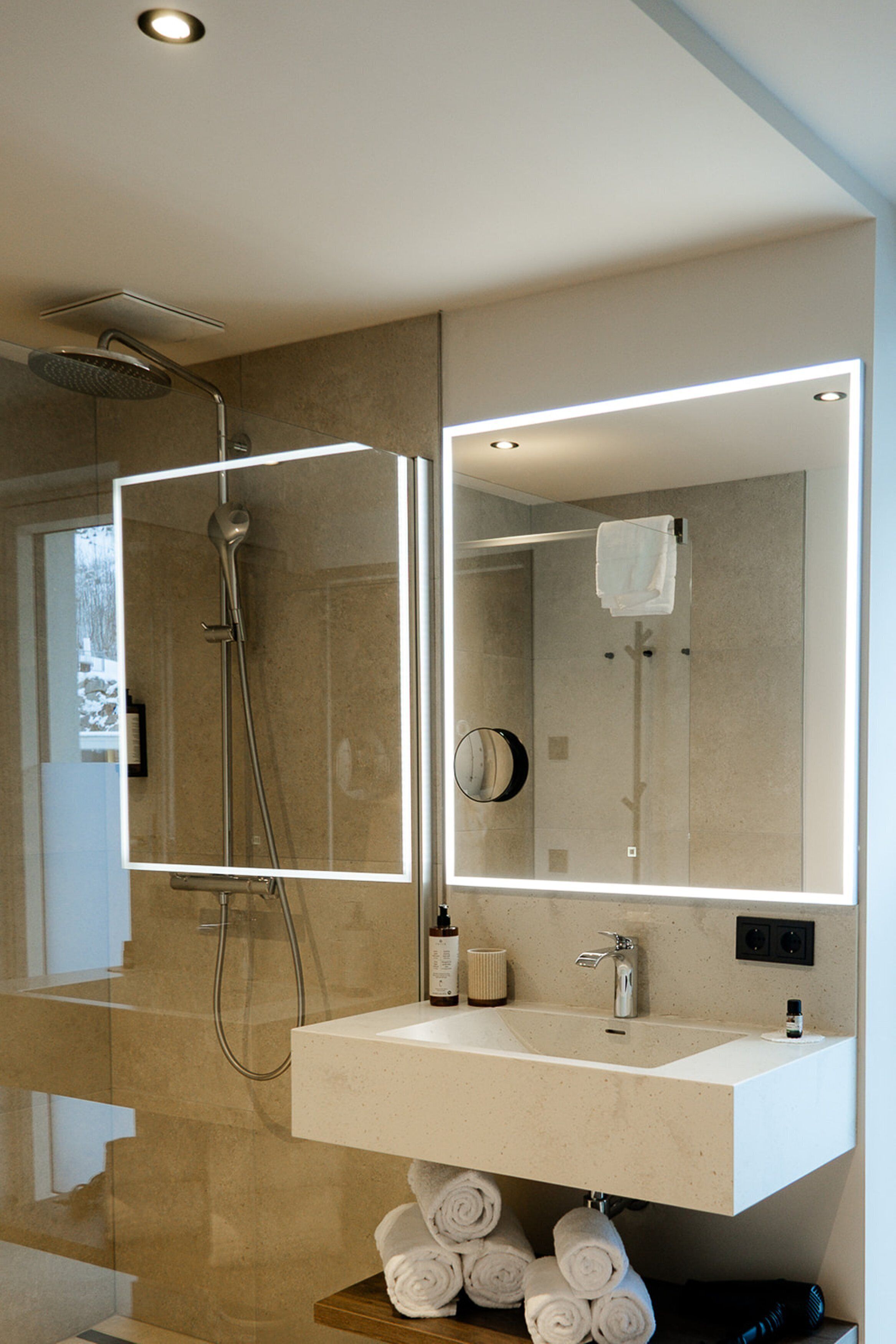 Un baño moderno con ducha envuelta en vidrio, un lavabo blanco y un espejo grande con iluminación integrada.