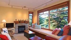 A cozy living room with a stone fireplace, a large window with a forest view, a comfortable sofa, and a television. - Bluff 9 townhouse with beautiful view (Whistler)