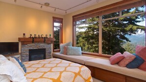 A cozy bedroom with a stone fireplace, a bed with patterned bedding, a bench with cushions, and a large window offering a scenic view. - Bluff 9 townhouse with beautiful view (Whistler)
