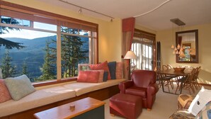 A cozy living room with a large window offering a mountain view, a dining area with a wooden table and chairs, and a seating area with a sofa and ottoman. - Bluff 9 townhouse with beautiful view (Whistler)
