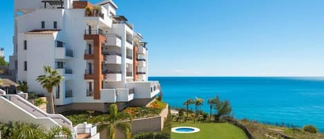 A modern multi-story hotel with a pool, surrounded by a well-maintained lawn and palm trees, overlooking a clear blue sea.