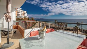 A balcony with a table set for two, a bottle of champagne, and a glass of water. The balcony overlooks a beach and a building.