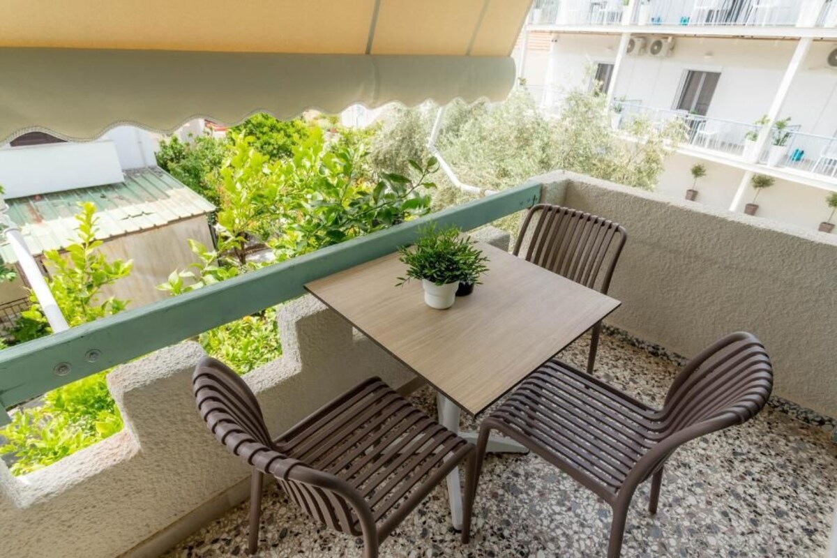 A balcony with a table and two chairs, a potted plant, and a view of greenery and buildings.