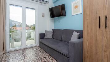 Une pièce avec un canapé gris, un téléviseur fixé au mur, une armoire en bois et une grande fenêtre donnant sur les bâtiments à l’extérieur.