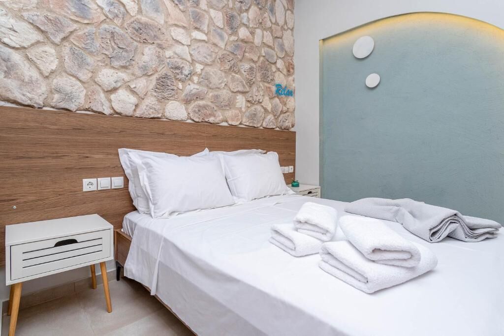 A neatly made bed with white linens and pillows, a stone wall, and a nightstand with a lamp.