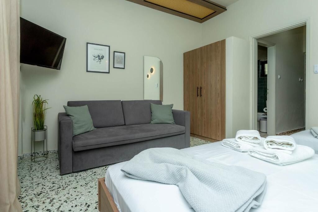 Una habitación de hotel moderna con un sofá gris, una cama con ropa blanca, un televisor montado en la pared y un armario de madera.