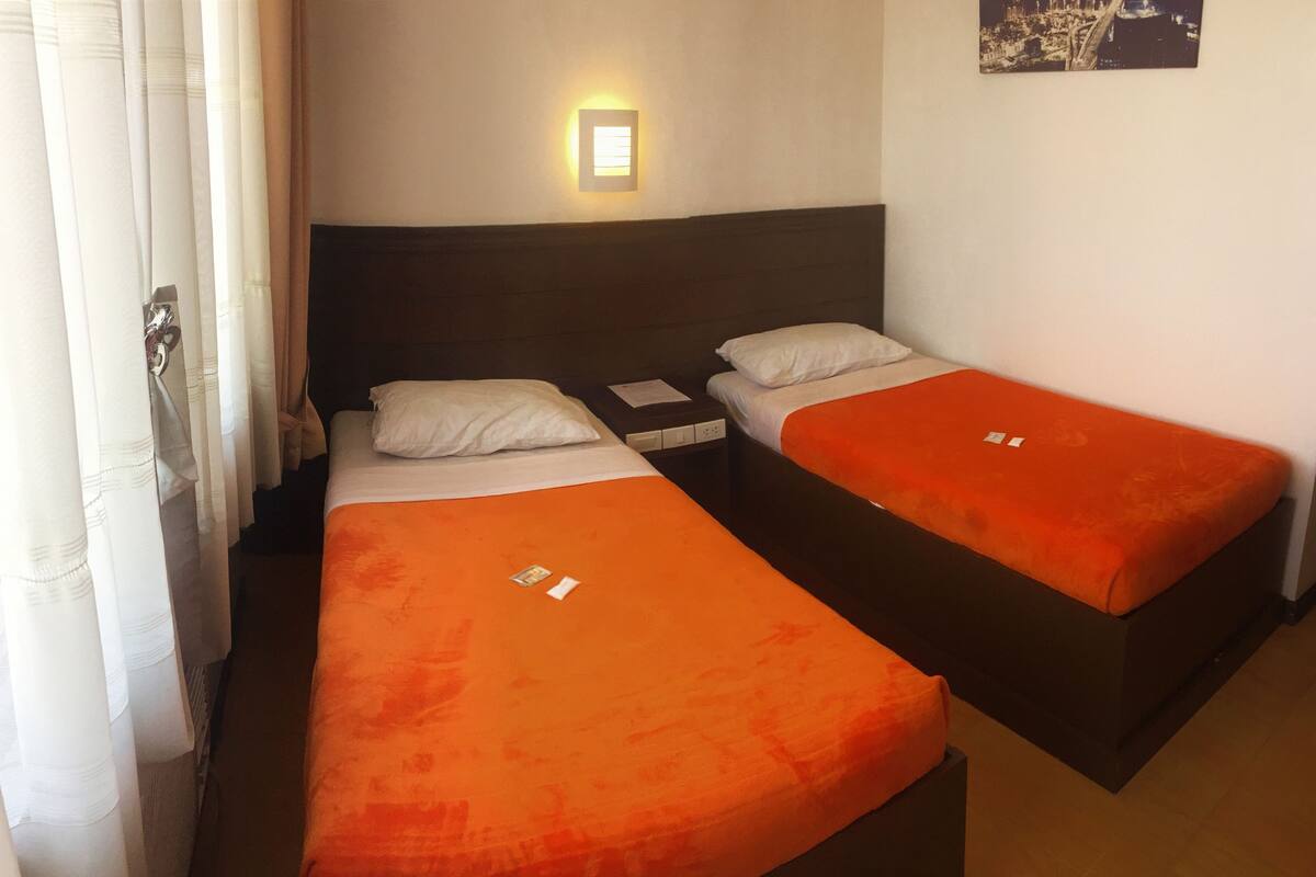 A hotel room with two single beds, a nightstand, a wall-mounted light, and a cityscape picture on the wall.
