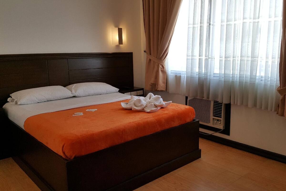 A hotel room with a bed, a wall-mounted air conditioner, and a window with curtains.