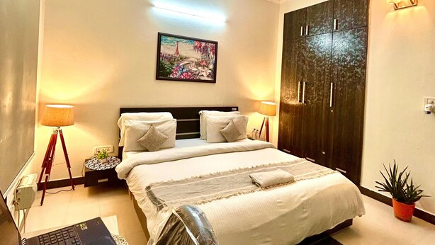 Une chambre d’hôtel avec un lit, un ventilateur de plafond, un bureau avec un ordinateur portable, une table de chevet, une lampe, un tableau et une porte.