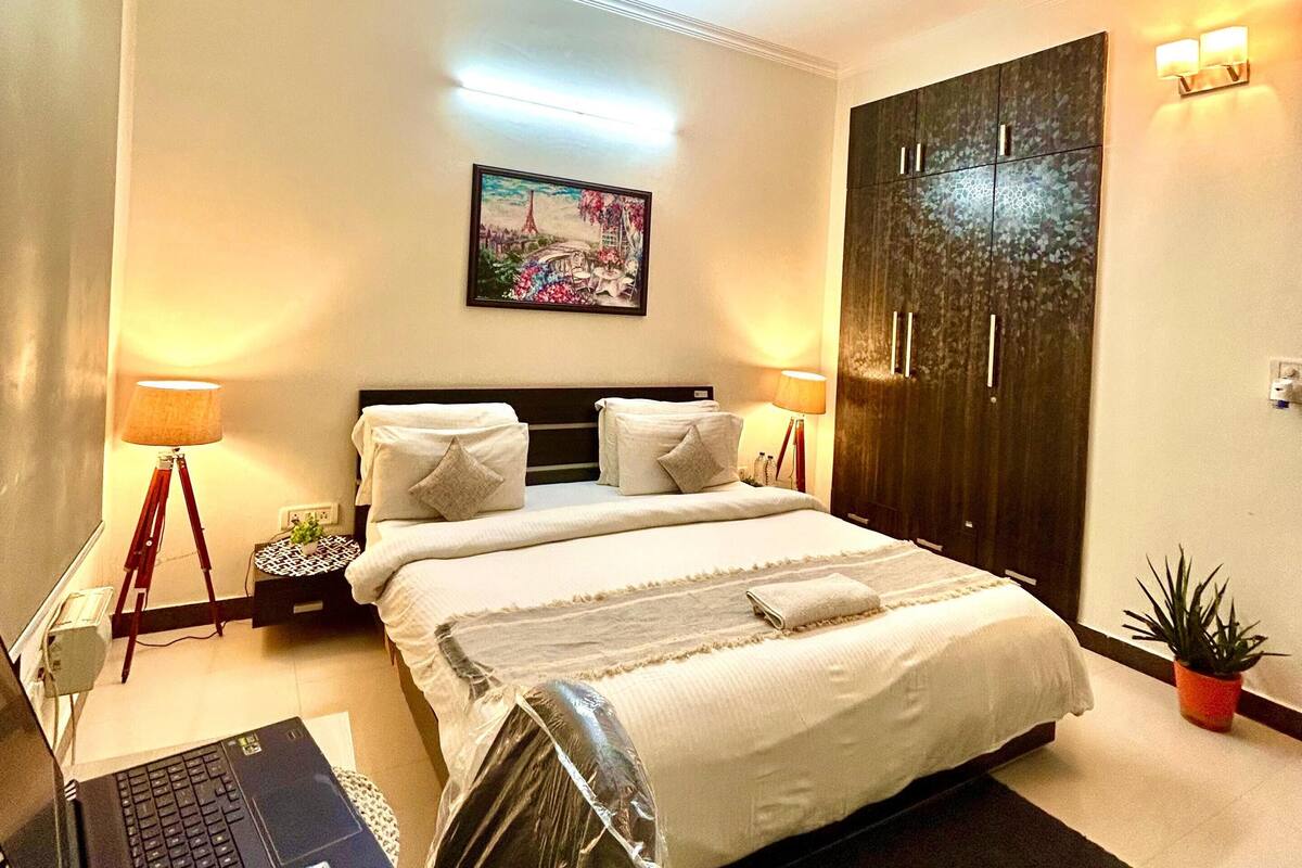 A hotel room with a bed, a ceiling fan, a desk with a laptop, a nightstand, a lamp, a painting, and a door.