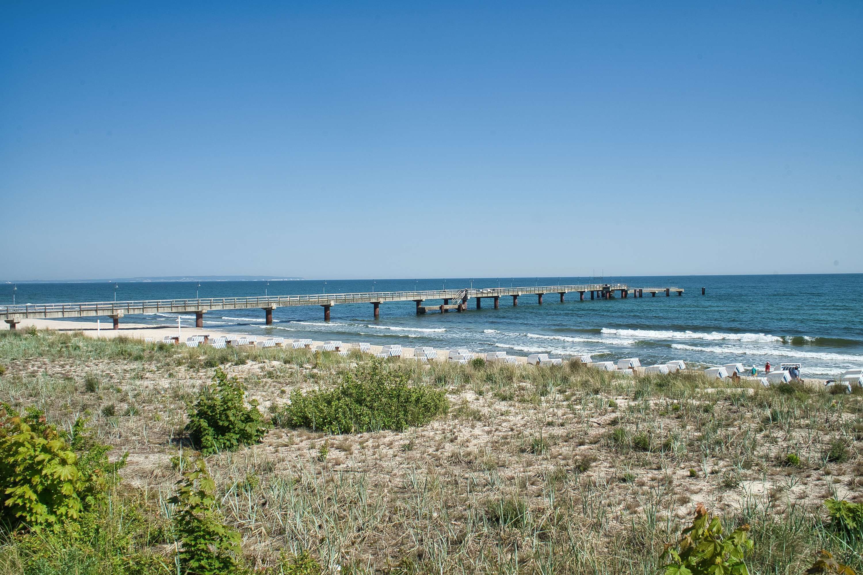 Vista costera con un muelle largo que se adentra en el mar, una playa de arena y vegetación escasa.