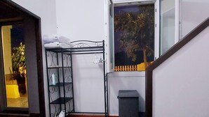 Ein Zimmer mit einem Holztisch, einem Metallregal mit Kleidung, einem Fenster mit Blick auf Bäume und einer Treppe.