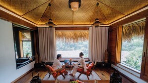 Une pièce dotée de planchers en bois, d’un plafond en chaume et de grandes fenêtres donnant sur un paysage verdoyant. On y trouve deux chaises en bois et une petite table sur laquelle est posé un panier de fruits.