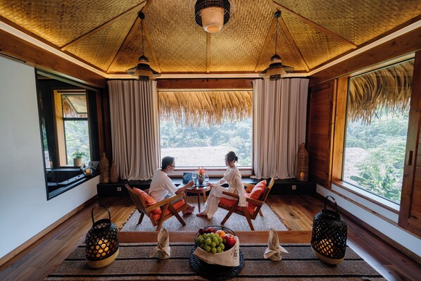 A room with wooden floors, a thatched ceiling, and large windows offering a view of greenery. There are two wooden chairs and a small table with a fruit bowl. - An Châng Retreat & Spa (Ha Giang 1)