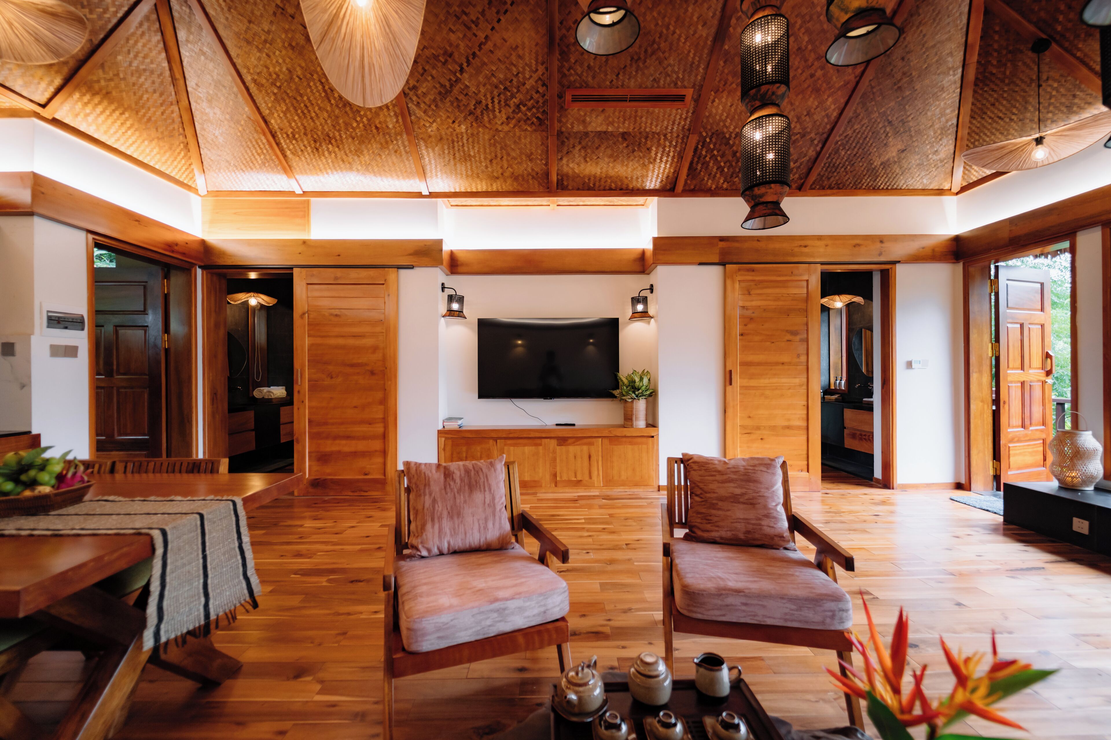 A spacious living area with wooden flooring, two cushioned chairs, a flat-screen TV, and a dining table with a basket of fruit.