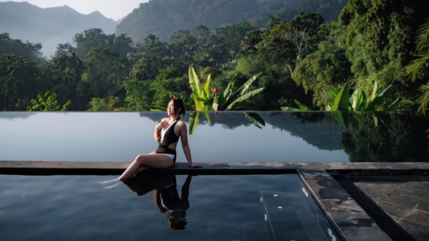 Une femme est assise au bord d’une piscine à débordement dont l’eau est parfaitement lisse, avec une végétation luxuriante et des montagnes en arrière-plan.
