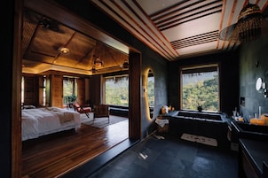 A room with a bed, a bench, a window with a view of the forest, and a bathroom with a sink and mirror.