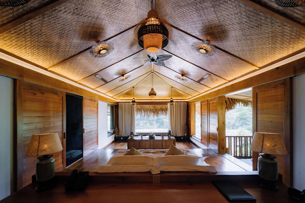 Una habitación con techo de madera, una cama con almohadas, un ventilador de techo y una ventana con cortinas.