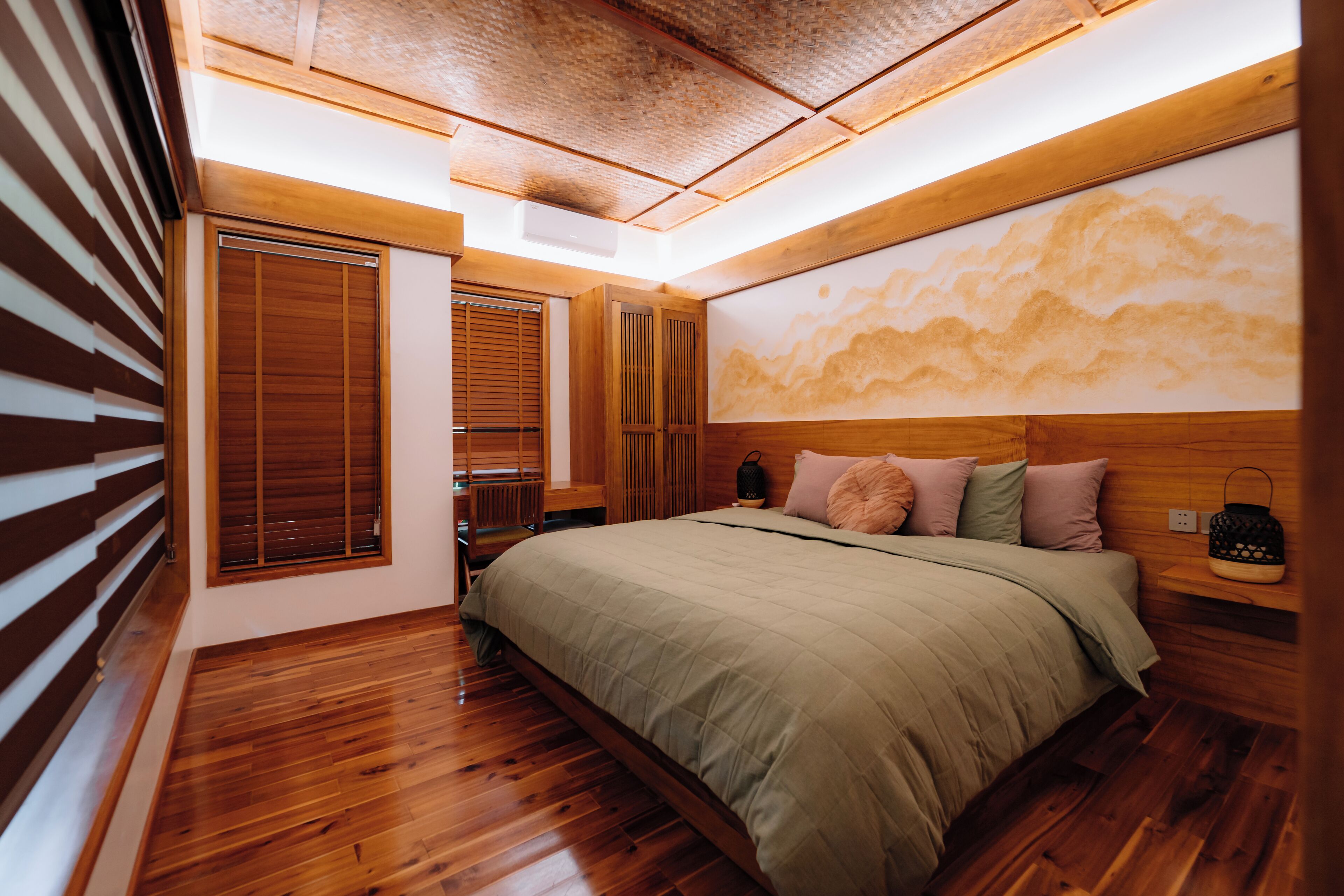 A bedroom with a wooden floor, a large bed with a green bedspread, wooden headboard, and a wall-mounted artwork.