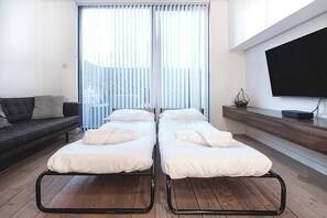 A modern room with two single beds, a dark sofa, a wooden floor, a large window with blinds, and a flat-screen TV mounted on the wall.