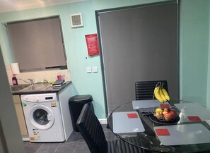 Une cuisine compacte équipée d’un lave-linge, d’une petite poubelle et d’un coin repas avec une table et des chaises en verre.